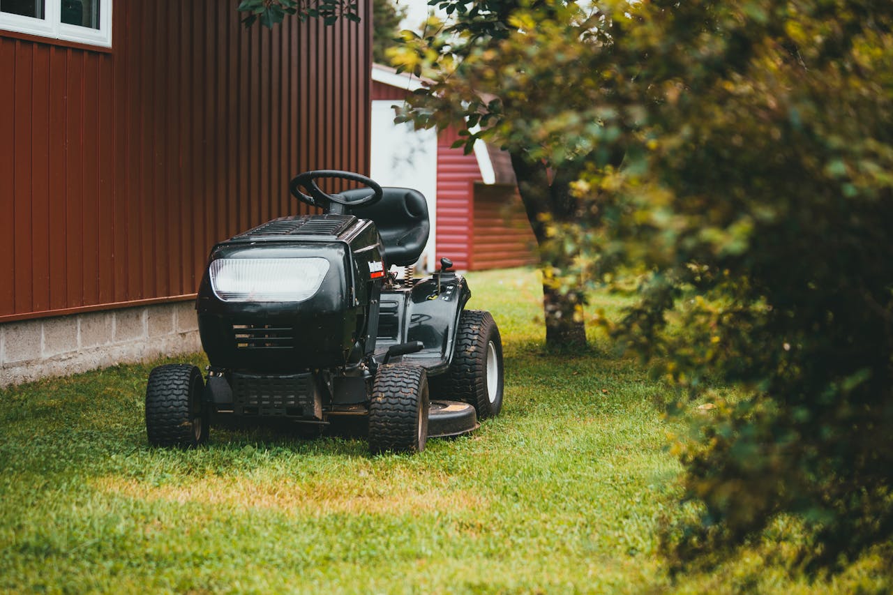 about-img A black ride-on lawnmower parked on green grass near a red barn during summer.
